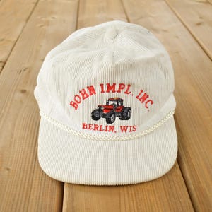 May include: White corduroy baseball cap with a red embroidered logo that says "BOHN IMPL. INC. BERLIN, WIS".