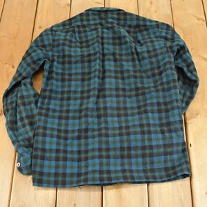 Op de afbeelding: Een lange mouwen overhemd met een klassiek ruitpatroon in teal, zwart en groen. Het overhemd is plat op een houten oppervlak gelegd, waarbij de achterkant van het kledingstuk te zien is. De stof lijkt zacht flanel.