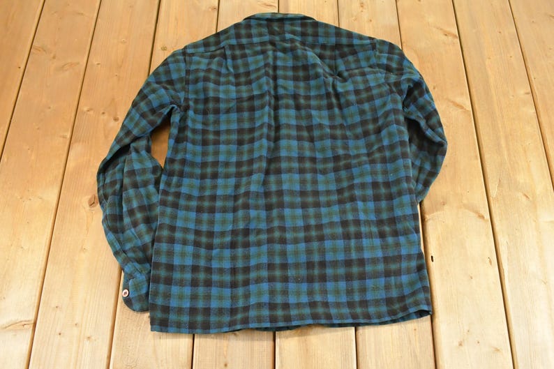 Op de afbeelding: Een lange mouwen overhemd met een klassiek ruitpatroon in teal, zwart en groen. Het overhemd is plat op een houten oppervlak gelegd, waarbij de achterkant van het kledingstuk te zien is. De stof lijkt zacht flanel.