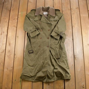 Pode incluir: Um casaco comprido de cor verde oliva, tipo trench coat, com gola de veludo cotelê castanho e fecho de dupla abotoadura. O casaco tem um cinto e dois bolsos.