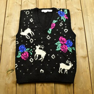 Op de afbeelding: Zwarte gebreide vest met een wit, roze, blauw en groen bloemen- en rendierpatroon.