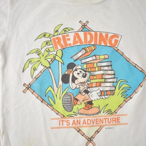 May include: White t-shirt with a cartoon image of Mickey Mouse carrying a stack of books. The image is surrounded by a blue and green tropical scene with palm trees and bamboo. The text "READING IT'S AN ADVENTURE" is printed below the image.