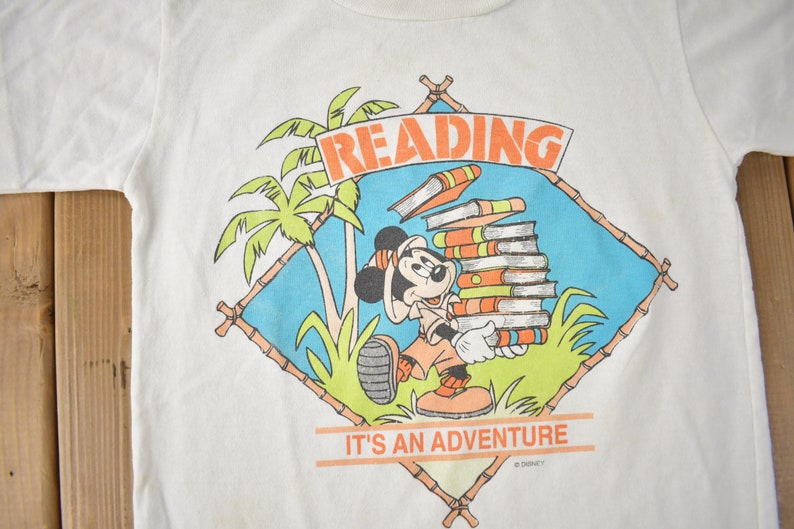 May include: White t-shirt with a cartoon image of Mickey Mouse carrying a stack of books. The image is surrounded by a blue and green tropical scene with palm trees and bamboo. The text "READING IT'S AN ADVENTURE" is printed below the image.