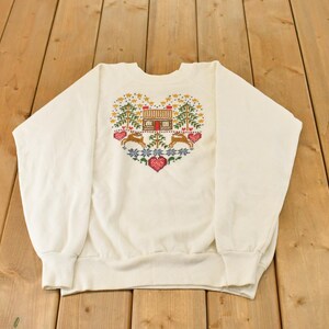 Puede incluir: Una sudadera de cuello redondo en color blanco roto con un gráfico central en forma de corazón. El diseño incluye una pequeña cabaña, dos ciervos, árboles de hoja perenne y varios patrones florales y geométricos en rojo, verde, azul y amarillo.