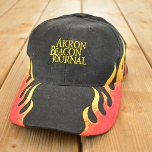 Puede incluir: Gorra de béisbol negra con el texto "AKRON BEACON JOURNAL" en amarillo. La visera presenta un diseño de llamas en rojo, naranja y amarillo. Es probable que el sombrero esté hecho de una tela duradera.