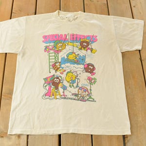 K&ouml;nnte beinhalten: Cremefarbenes T-Shirt mit einem farbenfrohen Aufdruck mit der Aufschrift "SPECIAL EFFECTS". Das Design zeigt Cartoonfiguren und Requisiten aus der Filmwelt, darunter eine Leiter, eine Kamera und ein Scheinwerfer. Das T-Shirt ist aus weichem Stoff.