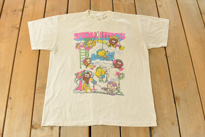 K&ouml;nnte beinhalten: Cremefarbenes T-Shirt mit einem farbenfrohen Aufdruck mit der Aufschrift "SPECIAL EFFECTS". Das Design zeigt Cartoonfiguren und Requisiten aus der Filmwelt, darunter eine Leiter, eine Kamera und ein Scheinwerfer. Das T-Shirt ist aus weichem Stoff.