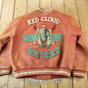 May include: A vintage red leather jacket with white and red striped trim. The back of the jacket features the text "RED CLOUD LAKOTA TRIBE AVIREX" with a bull skull and feathers graphic. The jacket has a button closure.