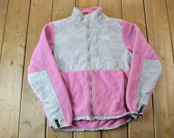 Puede incluir: Una chaqueta de forro polar rosa y gris con cierre de cremallera y un logotipo blanco de "The North Face" en el pecho.
