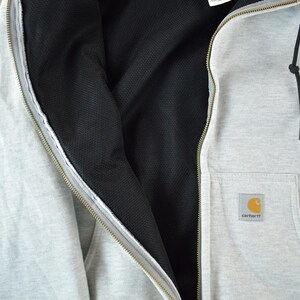 Puede incluir: Chaqueta con capucha gris claro con forro interior negro y bolsillo delantero. La chaqueta tiene una cremallera completa y un cord&oacute;n negro. Una peque&ntilde;a etiqueta cuadrada con un logotipo amarillo y naranja est&aacute; en el bolsillo.