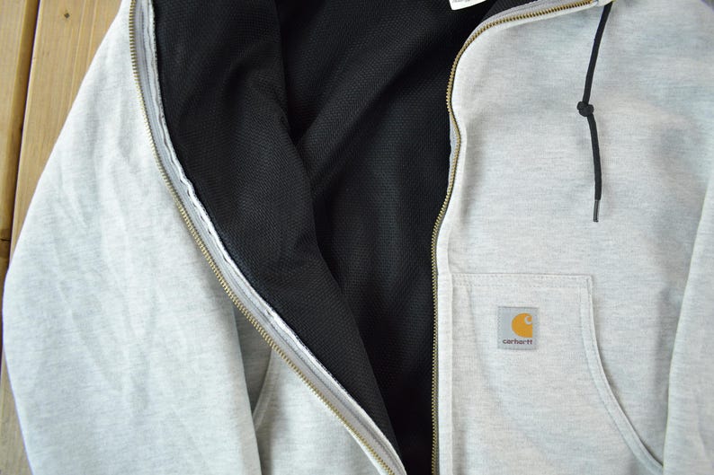 Puede incluir: Chaqueta con capucha gris claro con forro interior negro y bolsillo delantero. La chaqueta tiene una cremallera completa y un cord&oacute;n negro. Una peque&ntilde;a etiqueta cuadrada con un logotipo amarillo y naranja est&aacute; en el bolsillo.