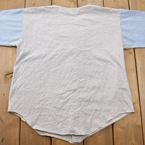 May include: A gray and light blue t-shirt with a baseball style design. The shirt has a light blue color on the sleeves and a gray color on the body.