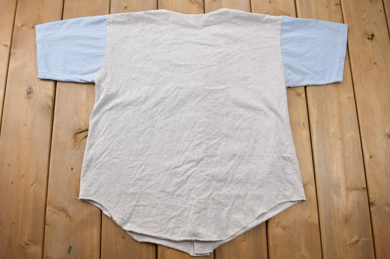 May include: A gray and light blue t-shirt with a baseball style design. The shirt has a light blue color on the sleeves and a gray color on the body.