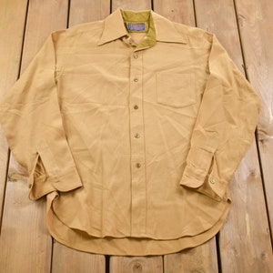 May include: A long-sleeved, tan button-down shirt with a single chest pocket. The shirt has a collar with a contrasting brown lining.