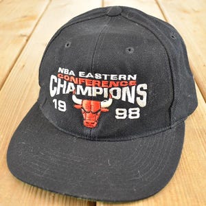 Può includere: Cappellino da baseball nero con la scritta "NBA Eastern Conference Champions 1998". Il cappellino presenta un logo di toro rosso e scritte bianche. È realizzato in tessuto resistente e ha una visiera curva.