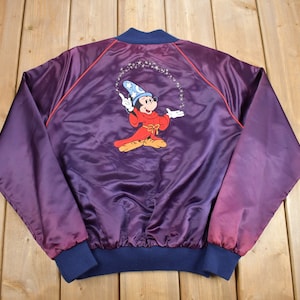 May include: A purple satin bomber jacket with a red and blue trim. The back of the jacket features an embroidered image of Mickey Mouse dressed as a sorcerer, with a blue hat and red robe, performing magic.
