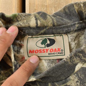 May include: A close-up of a camouflage pattern shirt collar with a label that reads "Mossy Oak Brand Camo". The shirt displays a mix of green, brown, and grey hues, common in hunting clothing.