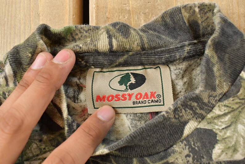 May include: A close-up of a camouflage pattern shirt collar with a label that reads "Mossy Oak Brand Camo". The shirt displays a mix of green, brown, and grey hues, common in hunting clothing.