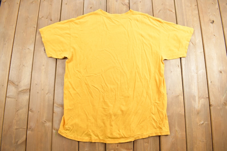 May include: A plain yellow t-shirt with short sleeves. The t-shirt is laid flat on a wooden surface.