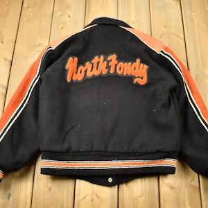 Può includere: Giacca da baseball nera con maniche a righe arancioni e bianche e una toppa di feltro sul retro che recita "North Fondy".