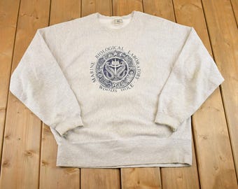 May include: Light gray sweatshirt with a crew neck. The front features a navy blue circular graphic with the text "MARINE BIOLOGICAL LABORATORY WOODS HOLE". The graphic includes a seahorse and other marine elements.