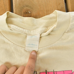 K&ouml;nnte beinhalten: Nahaufnahme eines cremefarbenen T-Shirts mit Rundhalsausschnitt. Das Shirt hat ein Etikett mit dem Markennamen "Little Dreams Originals". Das Shirt ist leicht zerknittert und scheint Vintage zu sein.
