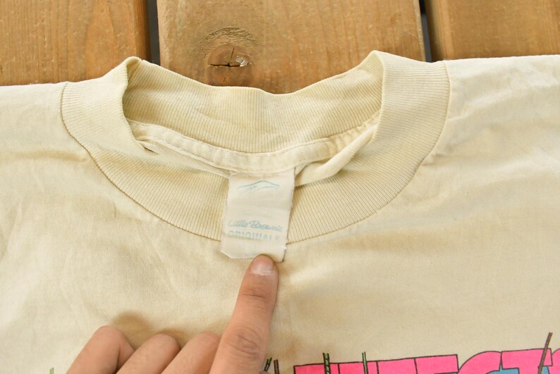 K&ouml;nnte beinhalten: Nahaufnahme eines cremefarbenen T-Shirts mit Rundhalsausschnitt. Das Shirt hat ein Etikett mit dem Markennamen "Little Dreams Originals". Das Shirt ist leicht zerknittert und scheint Vintage zu sein.