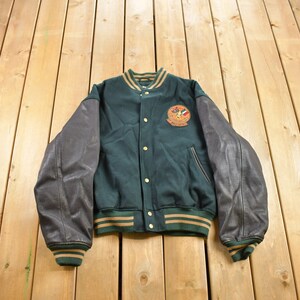 Può includere: Una giacca da baseball verde e marrone con maniche in pelle. La giacca ha una grande toppa ricamata sul petto sinistro con un logo e il testo "World Champions".