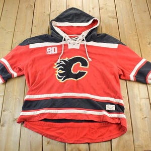 Puede incluir: Jersey de hockey rojo con capucha y mangas negras y blancas. La camiseta tiene el número 80 en el hombro izquierdo y un logotipo negro y amarillo en el pecho. Cordones blancos en el cuello.