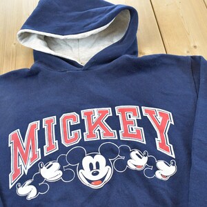 K&ouml;nnte beinhalten: Marineblauer Kapuzenpullover mit dem Wort "MICKEY" in Rot und Wei&szlig; sowie Mickey Mouses Gesicht und H&auml;nden. Die Kapuze hat ein hellgraues Futter.