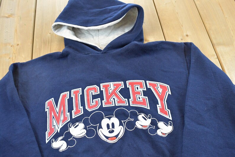 K&ouml;nnte beinhalten: Marineblauer Kapuzenpullover mit dem Wort "MICKEY" in Rot und Wei&szlig; sowie Mickey Mouses Gesicht und H&auml;nden. Die Kapuze hat ein hellgraues Futter.