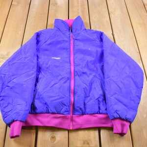 May include: A purple and pink fleece jacket with a zipper closure. The jacket has a stand-up collar and a logo on the left chest.
