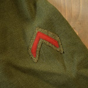 May include: A green military uniform with a red chevron patch sewn on the sleeve.
