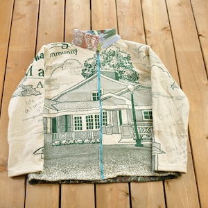May include: A cream-colored fleece jacket with a green and white print of a house and a street lamp. The jacket has a blue zipper and a high collar. The print includes the text "Community" and "Good Luck".