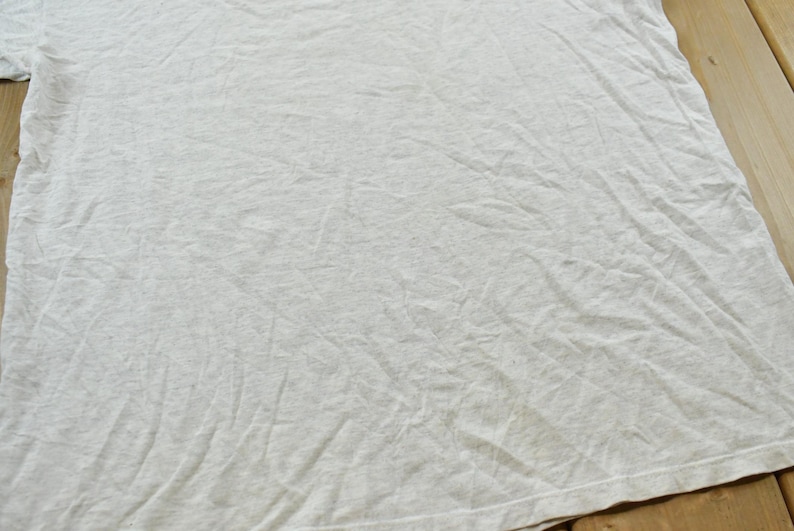 Op de afbeelding: Een lichtgrijs t-shirt met korte mouwen en een subtiele gespikkelde textuur door de hele stof. Het kledingstuk heeft een ronde halslijn en een standaard zoom, met een effen, onbedrukt oppervlak met zichtbare vouwen en kreukels in de stof.