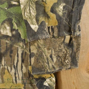 May include: Close-up of a long-sleeved camouflage shirt. The fabric features a pattern of green, brown, and tan, resembling a forest environment. The sleeve cuff is visible, showing the shirt's construction and material.