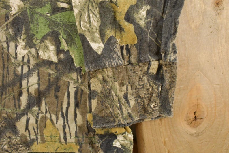 May include: Close-up of a long-sleeved camouflage shirt. The fabric features a pattern of green, brown, and tan, resembling a forest environment. The sleeve cuff is visible, showing the shirt's construction and material.