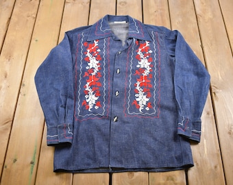 May include: A blue denim long-sleeve shirt with red and white embroidered floral designs on the front. The shirt has a button-up closure and a pointed collar.