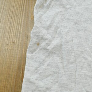Op de afbeelding: Een lichtgrijs gem&ecirc;leerd t-shirt met een zwarte grafische print aan de rechterkant. De stof heeft een zichtbare textuur en een klein bruin vlekje aan de linkerkant van het kledingstuk, nabij de rand van het mouwgedeelte.