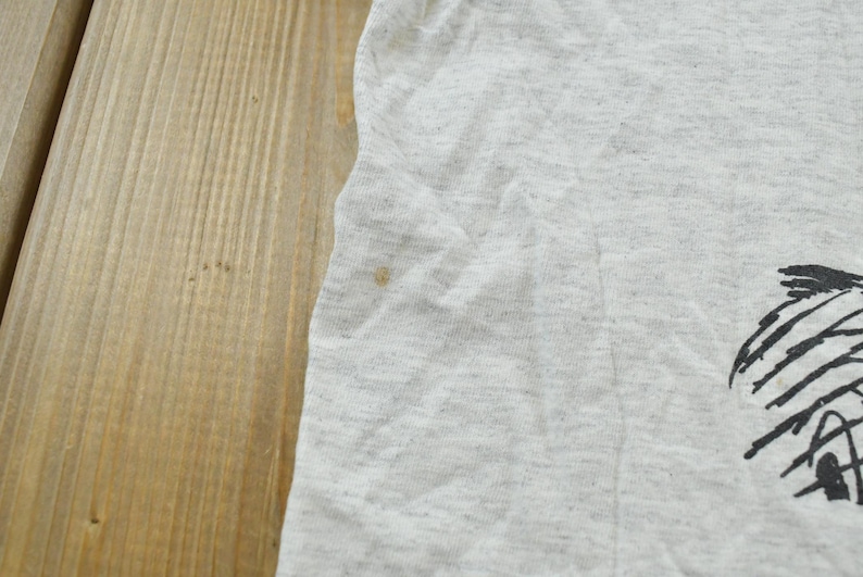 Op de afbeelding: Een lichtgrijs gem&ecirc;leerd t-shirt met een zwarte grafische print aan de rechterkant. De stof heeft een zichtbare textuur en een klein bruin vlekje aan de linkerkant van het kledingstuk, nabij de rand van het mouwgedeelte.