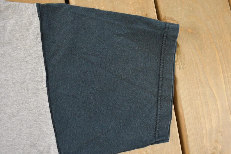 May include: Close-up of a two-tone t-shirt sleeve. The sleeve is divided into two sections: a light gray panel and a dark navy blue panel. The edge of the sleeve is hemmed with a visible seam.