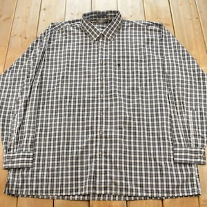 May include: A long-sleeve button-down shirt featuring a dark grey and white checkered pattern. The shirt includes a standard collar, a front button placket, and a small embroidered logo on the left chest area. The cuffs are finished with buttons.