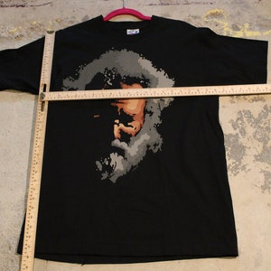 May include: Black t-shirt with a graphic print of a person's face in shades of gray and brown.