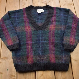 Può includere: Un maglione a collo a V a quadri blu scuro, verde e rosa. Il maglione è realizzato in un materiale morbido e soffice.