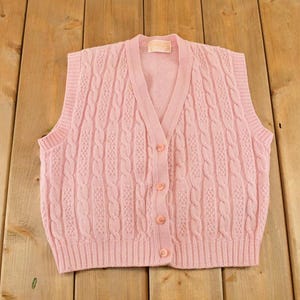 May include: A light pink sleeveless knit sweater vest featuring a V-neckline, a front button closure with four matching buttons, and a vertical cable knit pattern. The garment has ribbed trim along the neckline, armholes, and the bottom hem.