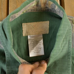 May include: Green button-up shirt with a camouflage pattern on the collar and shoulders. The shirt has a textured appearance and a tag with text. The shirt is XL/TG size.