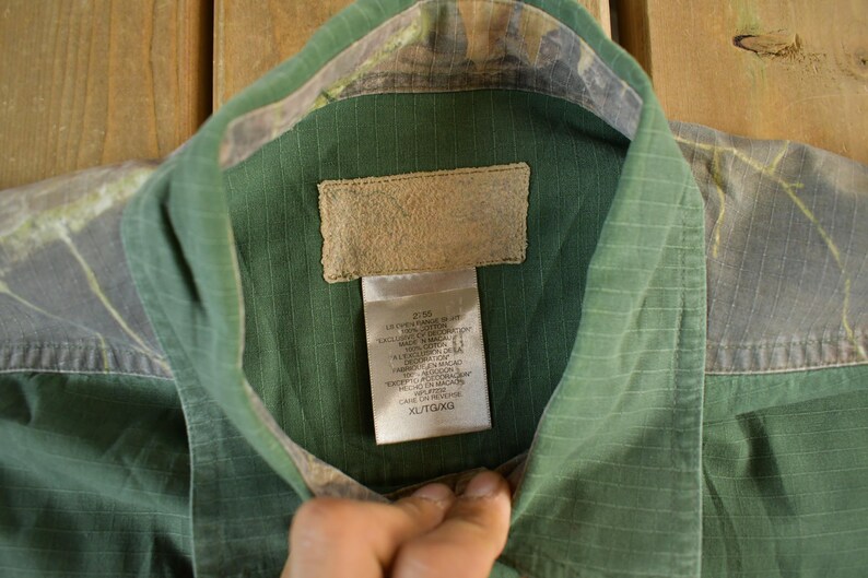 May include: Green button-up shirt with a camouflage pattern on the collar and shoulders. The shirt has a textured appearance and a tag with text. The shirt is XL/TG size.