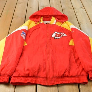 Puede incluir: Una chaqueta con capucha roja, amarilla y blanca que presenta el logotipo de los Kansas City Chiefs en el pecho y un parche con el escudo de la NFL en la manga derecha. La chaqueta tiene una cremallera frontal, mangas largas con puños elásticos y dobladillo acanalado.