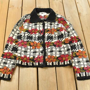 May include: A vintage, button-down sweater with a black and white houndstooth pattern. The sweater features a black collar and is adorned with floral designs in shades of red, orange, and yellow. The cuffs and hem are black.