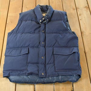 May include: A navy blue puffer vest with a stand-up collar and two front pockets. The vest has a button-up closure and is made of a quilted material. The vest is a classic outerwear piece.